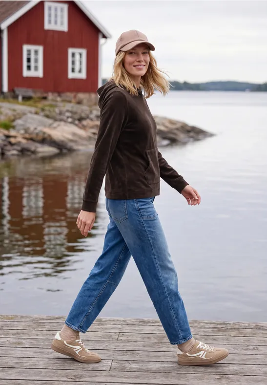Eine Frau mit Jeans und Jacke spaziert auf einem Steg am See vor einem roten Haus und lächelt.
