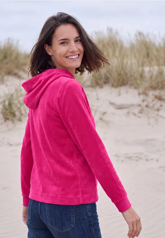Eine Frau in pinkem Hoodie und Jeans steht auf Sanddünen am Strand und lächelt zurück.