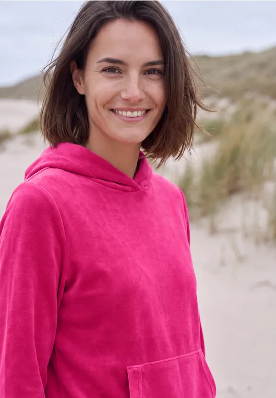 eine lächelnde Frau in pinkem Hoodie steht am Strand, im Hintergrund Dünen zu sehen.