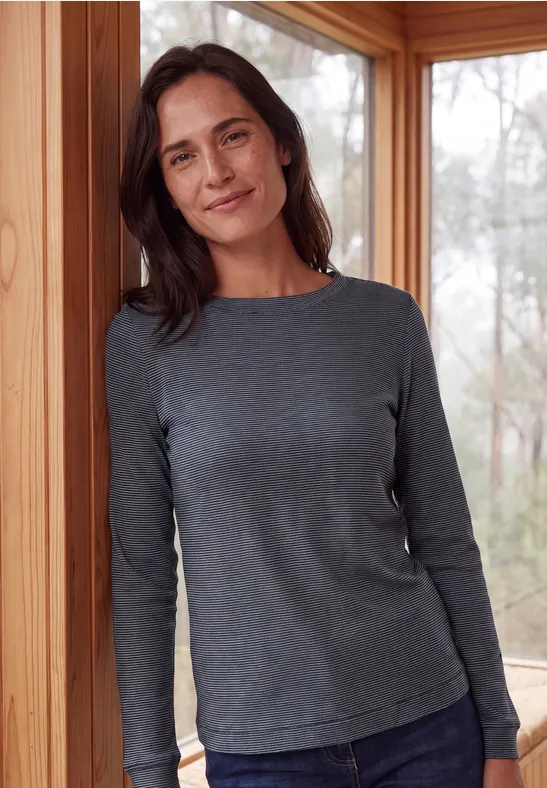 Eine Frau in grauem Langarmshirt lehnt lächelnd in einem hellen Holzhaus an einem Türrahmen.