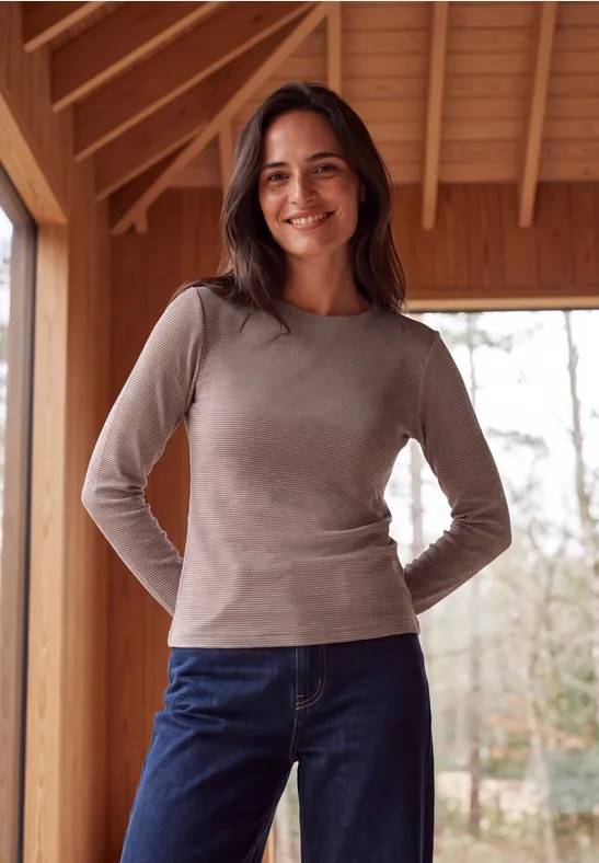 Eine lächelnde Frau in grauem Shirt und Jeans steht vor einer Holzwand mit Blick ins Freie.