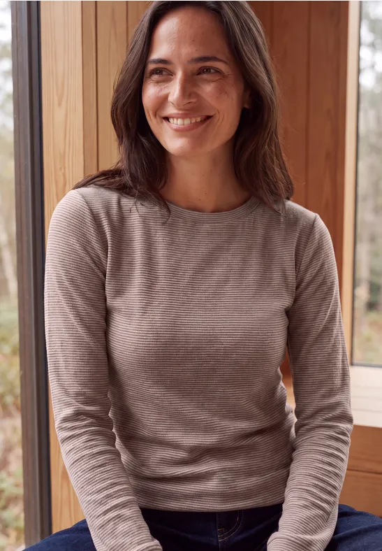 Eine lächelnde Frau im beigen Pullover sitzt entspannt vor einem Fenster in einer Holzhütte.