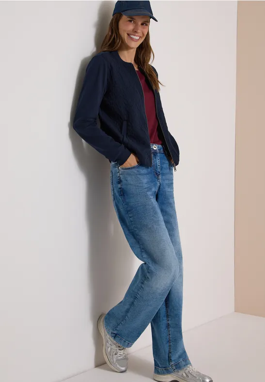 Frau lehnt an Wand in Jeans, dunkelblauem Cardigan, rotem Shirt, weißen Sneakern und Cap.