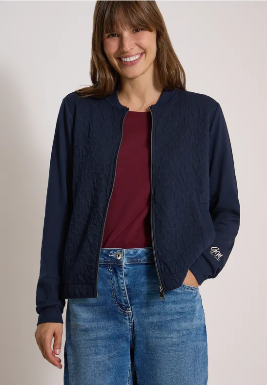 Junge Frau trägt blaue Jacke, bordeauxfarbenes Shirt und Jeans und lächelt.
