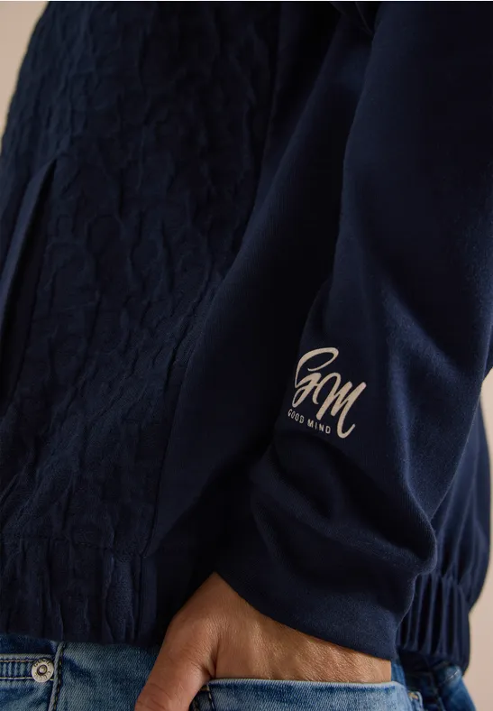 Nahaufnahme eines dunkelblauen Steppjackenärmels mit goldfarbenem HM-Logo am Bündchen und Hand in Jeans
