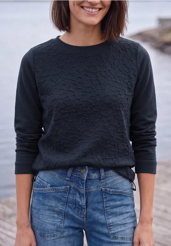 Eine lächelnde Frau in schwarzem Pullover und blauer Jeans vor einer Wasserkulisse.