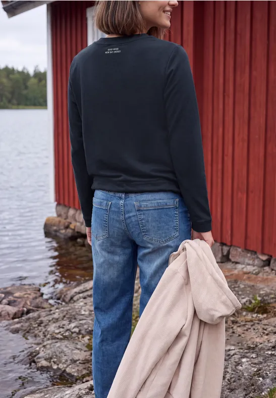 Eine Frau steht in Jeans und Pullover vor einem roten Holzhaus am See und hält einen beigen Mantel.