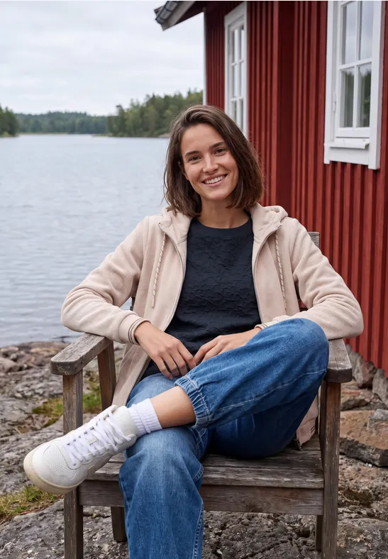 Eine lächelnde Frau in Jeans und Kapuzenjacke sitzt auf einem Holzstuhl vor einer roten Hütte am See.