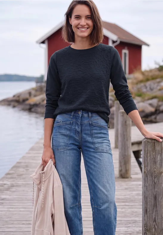 Frau im schwarzen Pullover und Jeans steht auf einem Steg vor einem roten Haus am Wasser und hält eine Jacke.