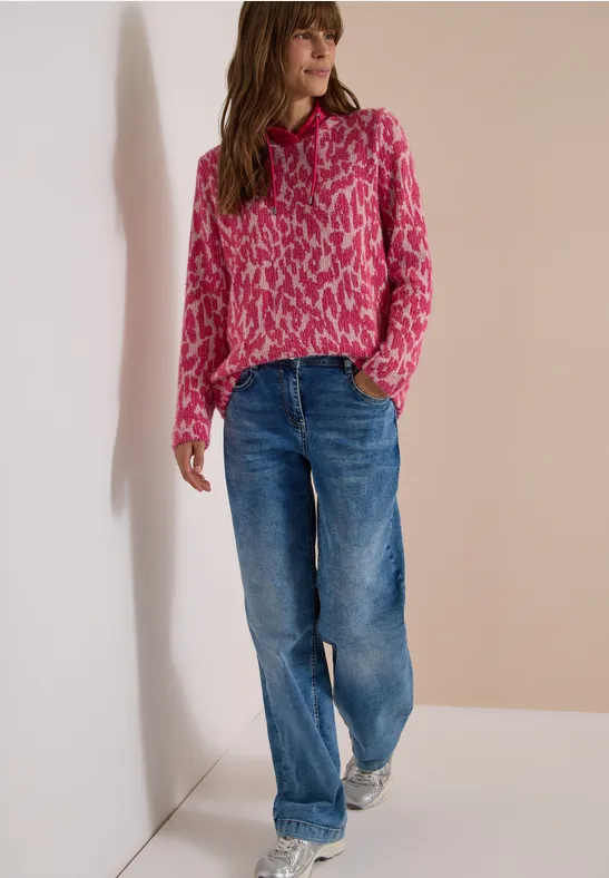 Frau steht an Wand, trägt pinkgemusterten Pullover und lockere blaue Jeans.