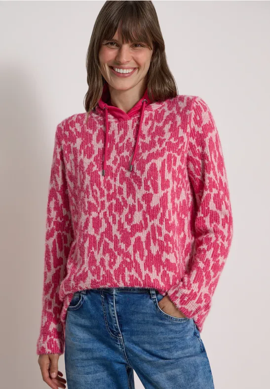 Frau mit pinkem, gemustertem Kapuzenpullover und Jeans, lächelnd vor neutralem Hintergrund.