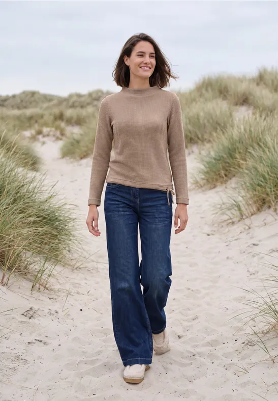 Une femme marche sur des dunes herbeuses au bord de la mer, en pull, jean et baskets blanches.