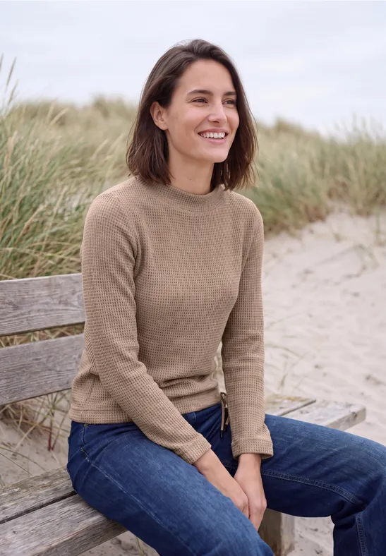 Femme assise sur un banc de plage, souriante en pull et jean, herbes et dunes en arrière-plan.