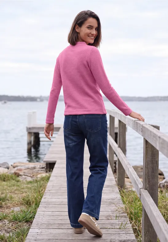 Frau in pinkem Pullover und Jeans steht auf einem Steg am Wasser und blickt zurück.