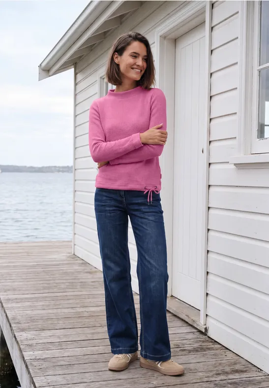 Frau in pinkem Pullover und Jeans steht auf Holzsteg vor weißem Haus am Wasser und lächelt.