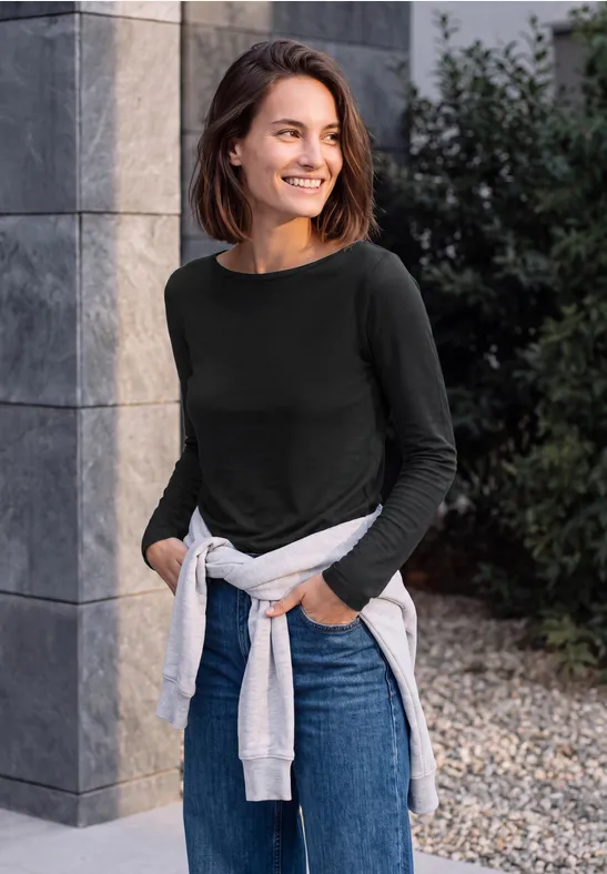 Lachende vrouw leunt tegen een grijze muur, draagt zwarte trui, jeans en licht sweatshirt om haar middel.