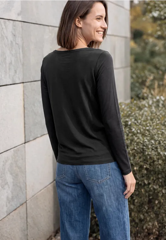 Vrouw in zwart shirt en blauwe spijkerbroek, deels van achteren gezien bij stenen muur en struiken.