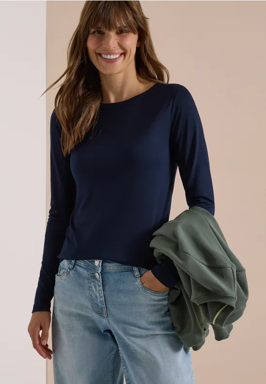 Lächelnde Frau in dunkelblauem Langarmshirt und Jeans, hält eine grüne Jacke und steckt eine Hand in die Hosentasche.