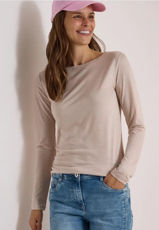 Eine lächelnde Frau in beigem Langarmshirt, Jeans und rosa Cap vor hellem Hintergrund.