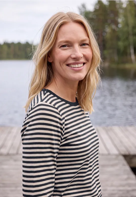 Eine lächelnde blonde Frau in gestreiftem Shirt steht an einem Steg am See, umgeben von Wald.