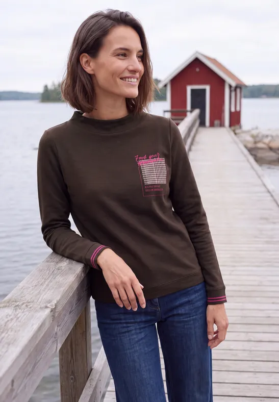 Eine Frau lehnt lächelnd an einem Holzgeländer auf einem Steg vor einem roten Häuschen am See.