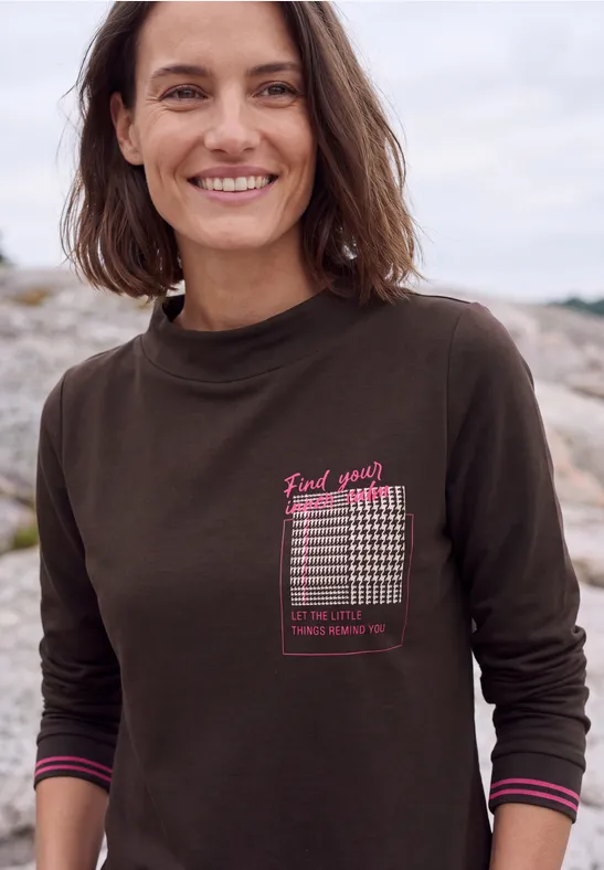 Frau in dunkelgrünem Langarmshirt mit Print, lächelt vor felsiger Küstenkulisse.