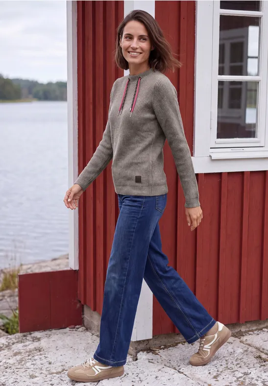 Eine Frau geht lächelnd an einem roten Haus am See entlang, trägt Jeans und Pullover.
