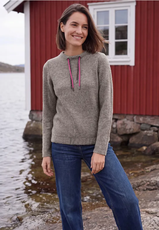 Frau in Pullover und Jeans steht lächelnd vor rotem Holzhaus am Wasser.