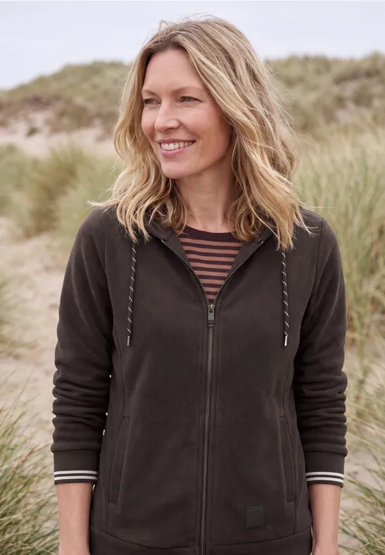 Une femme souriante en veste à capuche se tient devant des dunes de sable et des herbes de plage.