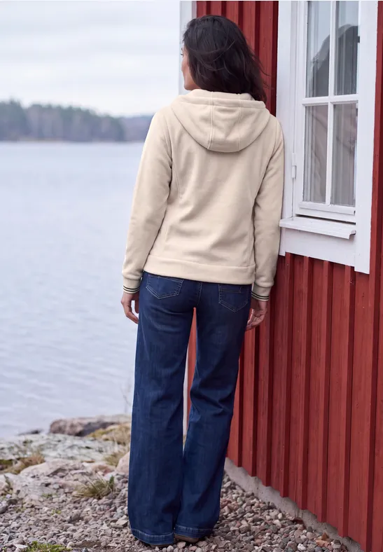 Person mit beiger Kapuzenjacke und blauer Jeans steht mit Blick auf einen See neben rotem Holzhäuschen.