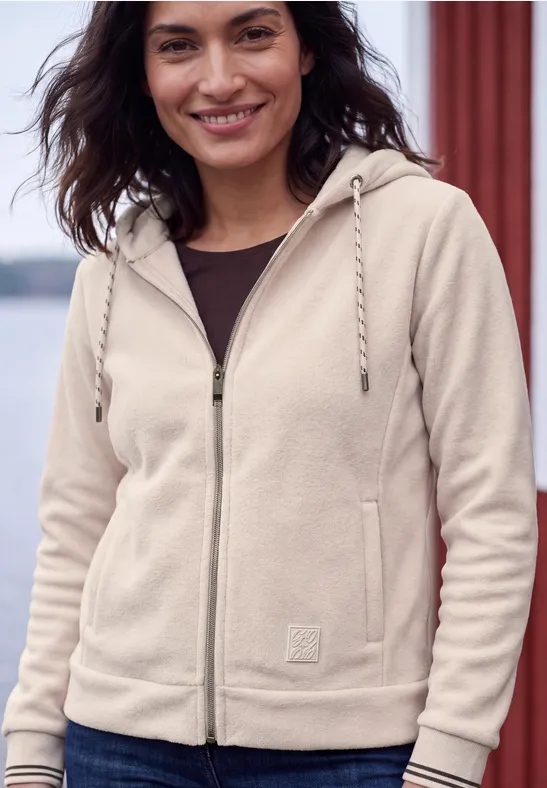 Eine lächelnde Frau trägt einen beigen Reißverschluss-Hoodie mit Kordelzug und Taschen.
