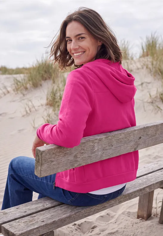 Eine lächelnde Frau in pinker Jacke sitzt auf einer Holbank am Dünenstrand.