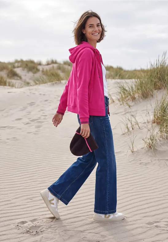 Frau in pinkem Hoodie, blauen Jeans und weißen Sneakern spaziert lächelnd durch Dünen und hält einen Hut.