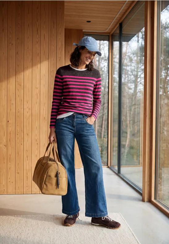 Vrouw in roze-blauw gestreepte trui, wijd jeans, blauwe pet en tas staat in een lichte houten gang met ramen.