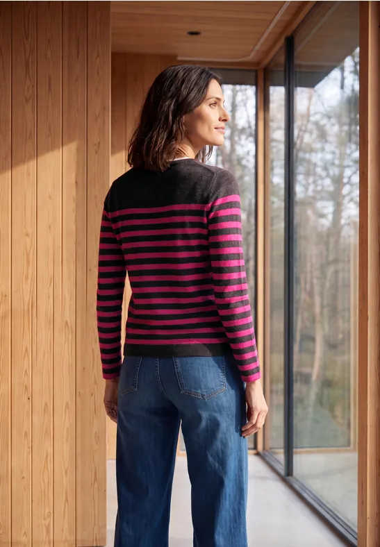 Een vrouw in een gestreepte trui staat binnen bij een raam en kijkt naar het bos