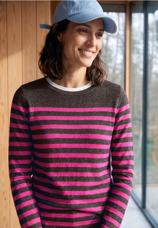 Vrouw met blauwe pet en grijs-fuchsia gestreepte trui, glimlachend voor een raam.