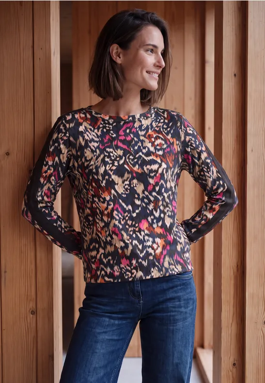 Een glimlachende vrouw in kleurrijke, gemêleerde trui en jeans voor een houten wand.