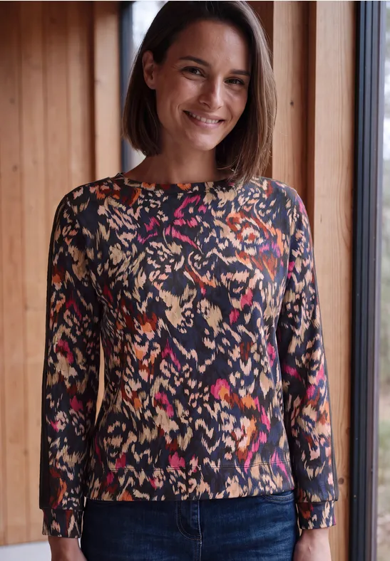 Een glimlachende vrouw staat bij een raam in een kleurrijk, patroonshirt met lange mouwen.