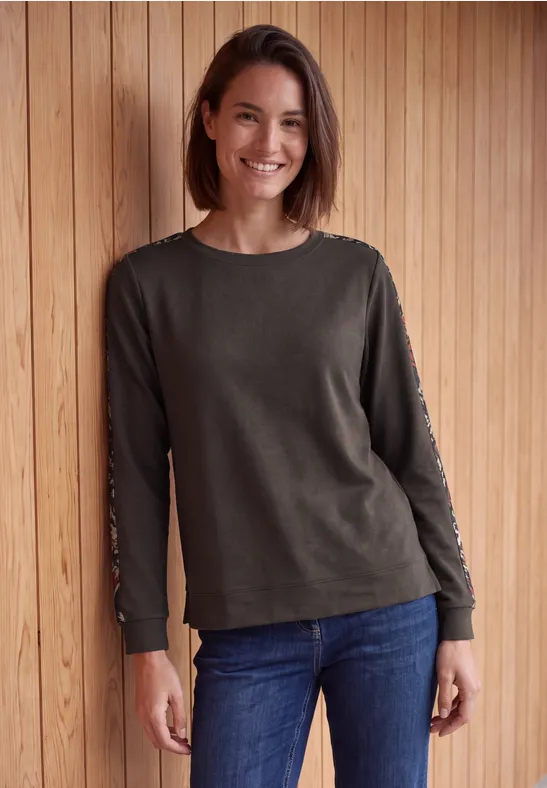 Eine lächelnde Frau mit schulterlangem Haar, grauem Langarmshirt und blauer Jeans vor Holzwand.