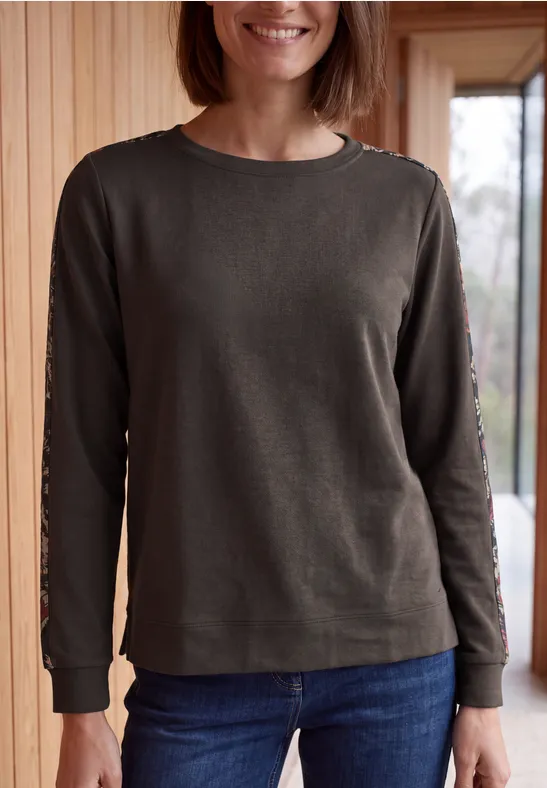 Frau mit grünem Langarmshirt und Jeans steht lächelnd vor einer Holzpaneelwand.