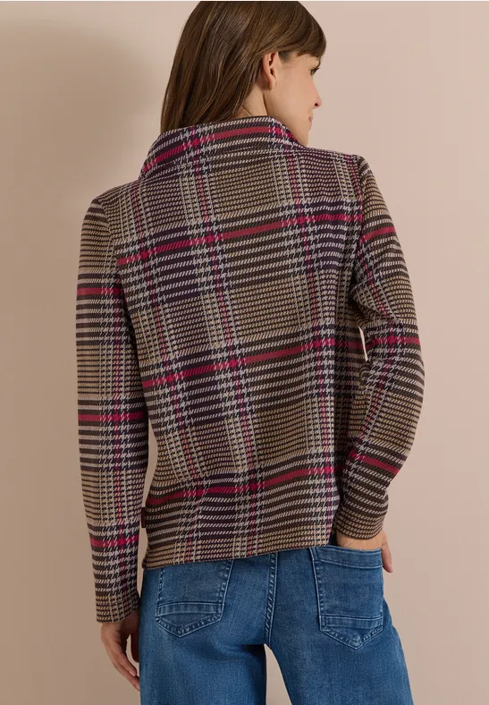 Frau von hinten in karierter Jacke mit roten Streifen und blauen Jeans vor beigem Hintergrund.