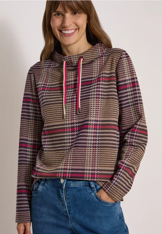 Lächelnde Frau im karierten Braun-Pink-Hoodie und blauen Jeans vor weißem Hintergrund.