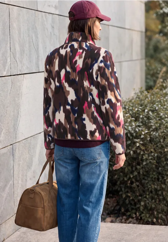 Vrouw met rode pet, dessinjas en blauwe jeans draagt een bruine tas voor een muur en struiken.