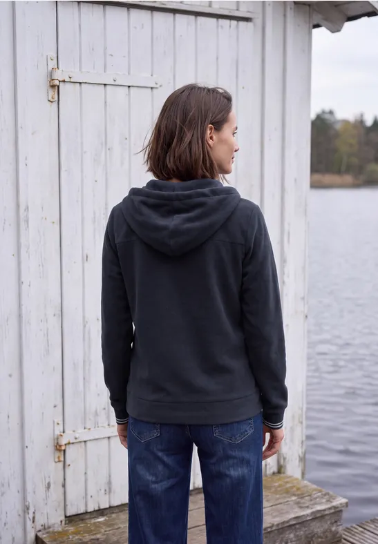 Rückansicht einer Person in dunkelgrauer Kapuzenjacke vor einem weißen Holzhaus am Wasser.