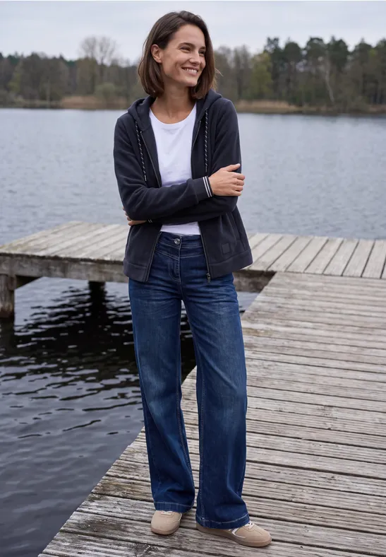 Frau lächelnd in Jeans und Cardigan steht auf einem Steg am ruhigen See mit Wald im Hintergrund.