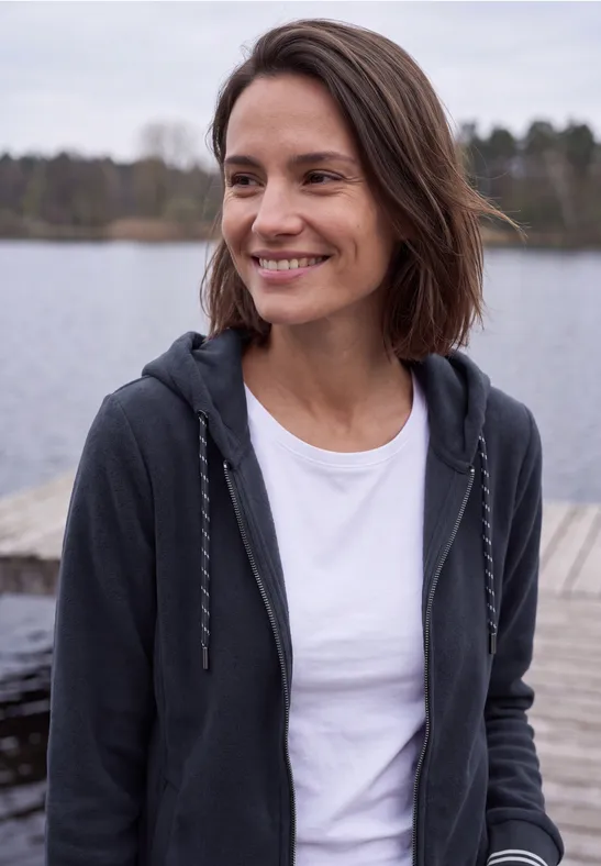 Eine lächelnde Frau im weißen T-Shirt und Hoodie steht auf einem Steg am See, im Hintergrund Wald.