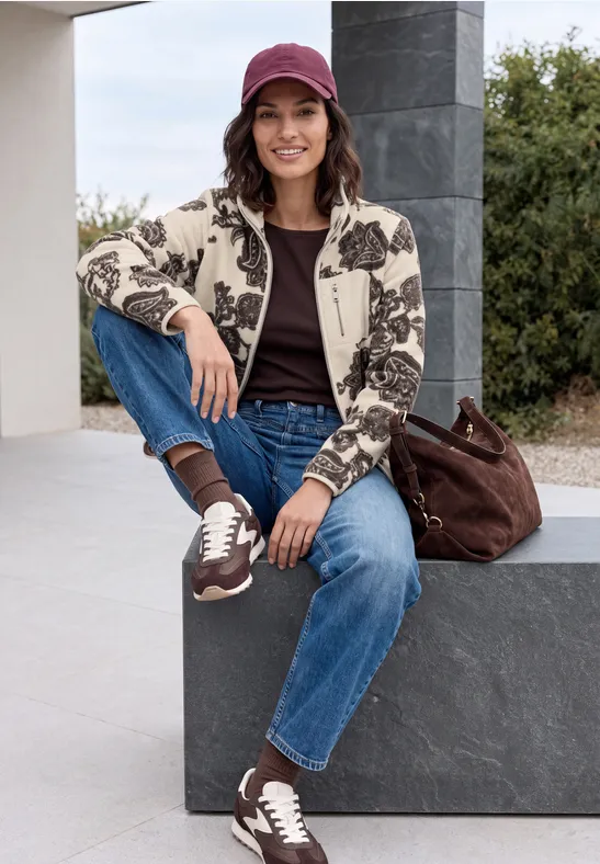 Eine Frau in Jeans, bedruckter Strickjacke und Cap sitzt lächelnd auf Steinbank.