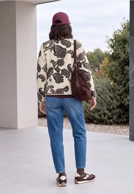 Person von hinten in gemusterter Jacke, blauer Jeans, Sneakern und roter Kappe mit brauner Umhängetasche draußen.