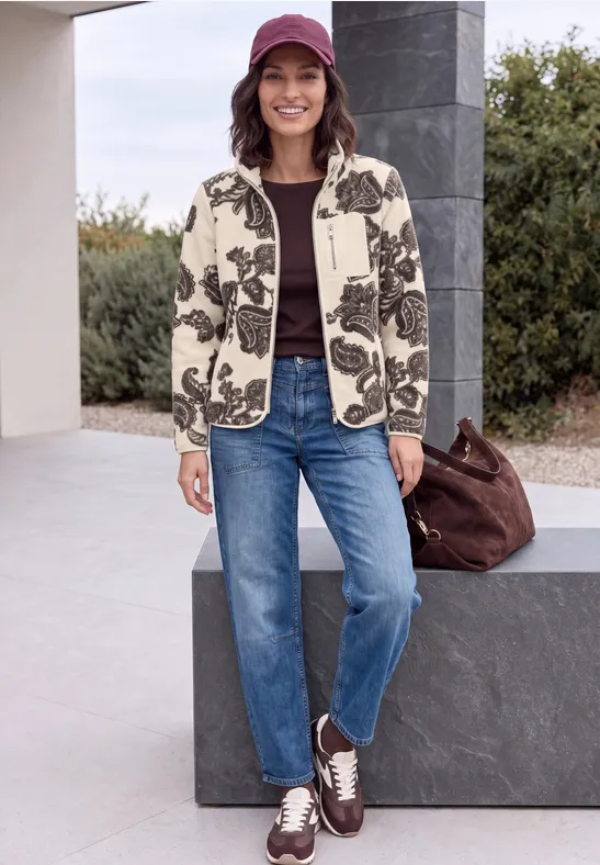 Frau in Blumenjacke, Jeans, Sneakern und Cap steht lächelnd vor moderner Hausfassade.
