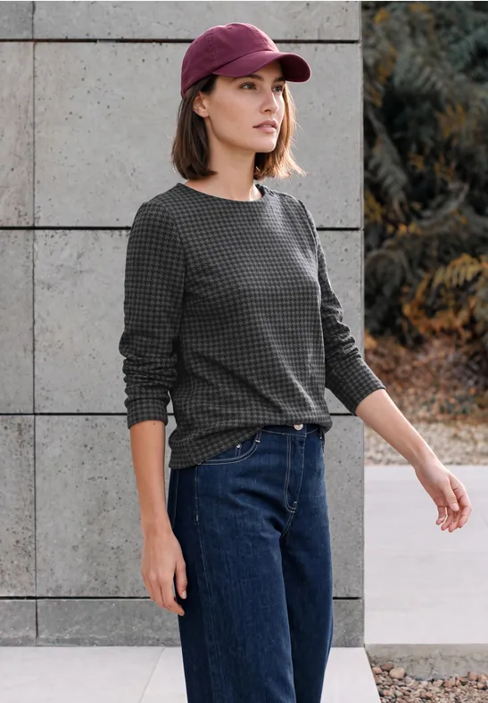 Frau mit rosafarbenem Cap, grauem Pullover und Jeans steht vor Wand und Pflanzen.